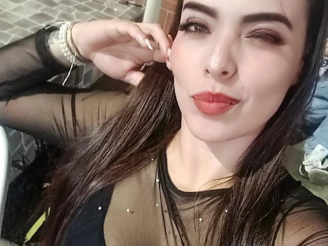 LorenDolce has long brown hair, winking with one eye, wearing sheer black top, red lipstick, and pearl bracelet, smiling slightly, touching her ear with her right hand.