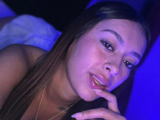 DamiAmbani with long brown hair, blue-lit face, slightly open mouth, finger touching lips, glossy pink lips, subtle tattoo on chest, wearing a gold necklace, looking seductive.