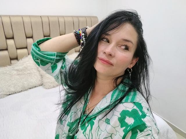Alaishka with long black hair, green floral shirt, hand in hair, smiling, wearing earrings and bracelets, on white bed with beige headboard. Her tits are small and she's cute.