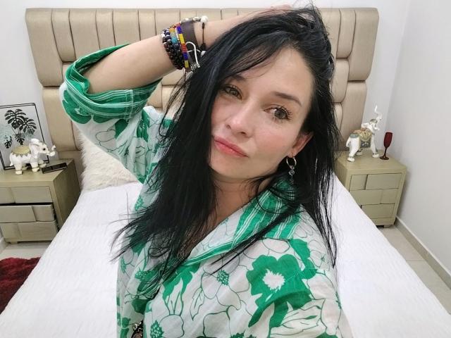 Alaishka, with long black hair, green floral robe, and bracelets, raises her hand, showing off her tits, and has a slight smile, sitting on a bed.