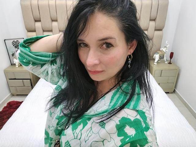 Alaishka with green floral shirt, black hair, pink cheeks, green earrings, right hand in hair, white bed, beige headboard, red rug, and white walls in the background.