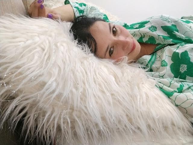 Alaishka with purple nails, lying on a fluffy white pillow, wearing a green floral shirt, looking up with a slight smile, dark hair, and green eyes.