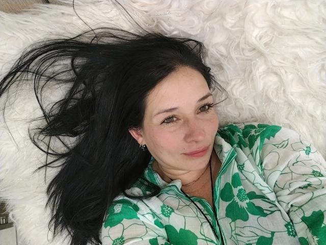 Alaishka with black hair lying on white fluffy pillow, wearing green floral pajamas, smiling slightly, small earrings, green eyes, fair skin, casual, relaxed, cute, soft lighting.