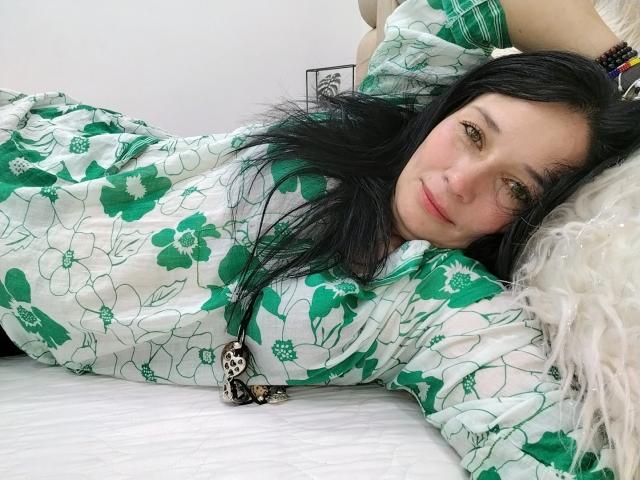 Alaishka with black hair, green floral pajamas, lying on white bed, smiling, arm raised, wearing black monkey necklace, white fluffy pillow, relaxed, attractive, cute, soft lighting.