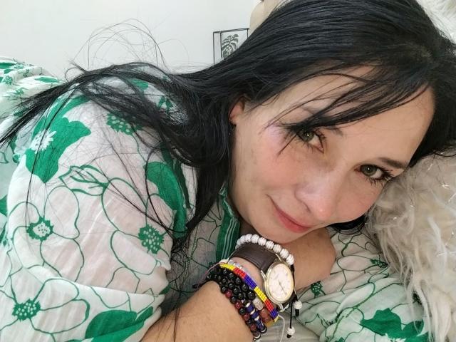 Alaishka with long black hair, green eyes, and light skin, lying on a bed, wearing a green floral shirt, and colorful wristbands, smiling gently at the camera.