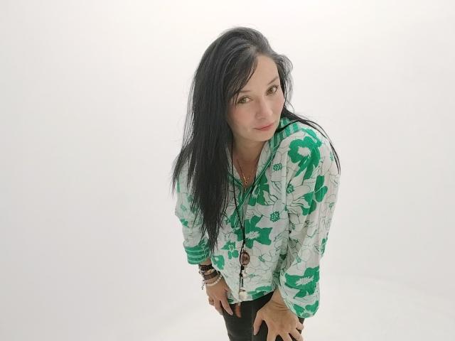Alaishka with long black hair, green floral shirt, leaning forward, hands on knees, white background, slightly smiling, green eyes, wearing black pants, necklace with brown beads.