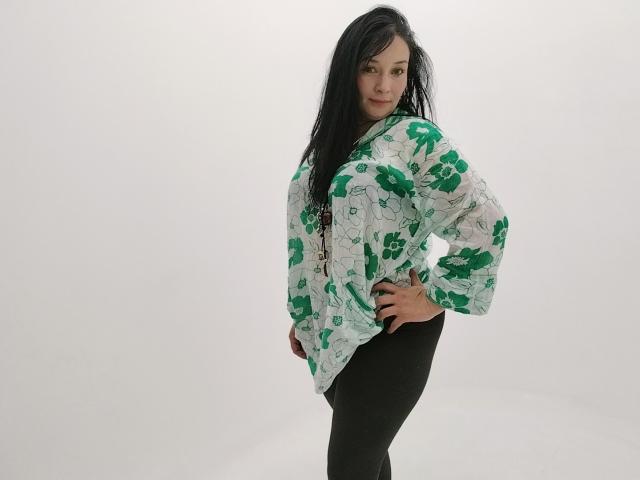 Alaishka with long black hair, green and white floral shirt, black pants, hands on hips, looking over shoulder, slight smile, white background, standing, confident, casual.