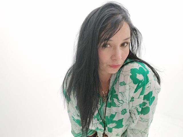 Alaishka with long black hair, green floral shirt, looking over shoulder, slight smile, white background, light skin, subtle makeup, small gold necklace, relaxed, attractive.