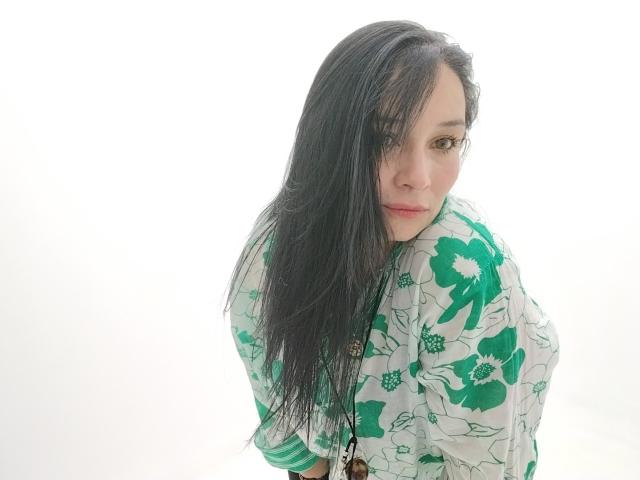 Alaishka with long black hair, green floral shirt, looking over shoulder, soft white background, slight smile, green eyes, light skin, subtle makeup, loose hair covering right eye.