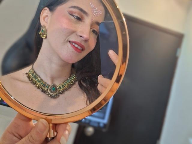 JalimaMona with dark hair, red lipstick, and green jeweled necklace, smiling at herself in a round mirror, holding a phone. She's wearing a decorative forehead dot.