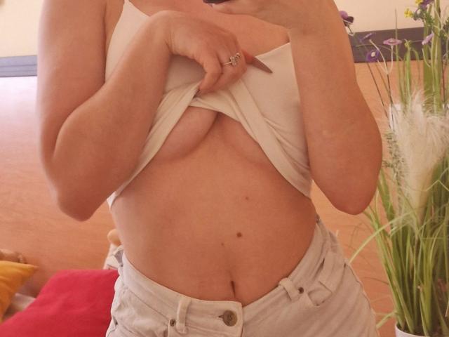 Jumiera lifting a white tank top to expose her bare, medium-sized boobs, with a ring on her right hand and white jeans unbuttoned, showing her flat stomach.