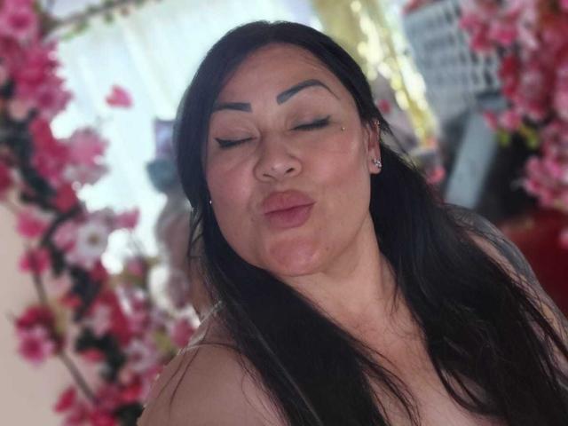 KittyHotanal with black hair, thick eyebrows, closed eyes, puckered lips, and small earrings, surrounded by pink flowers, looking cute and sexy.
