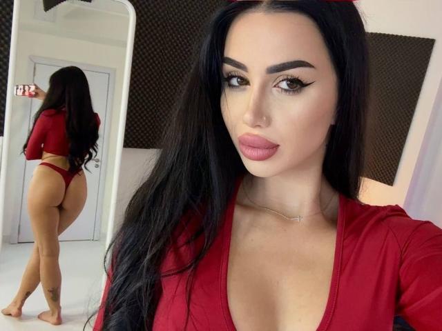 JennaDay with long black hair, red top, and thong, showing her ass in mirror selfie, full tits, sexy pink lipstick, tattoo on right calf, looking at camera.