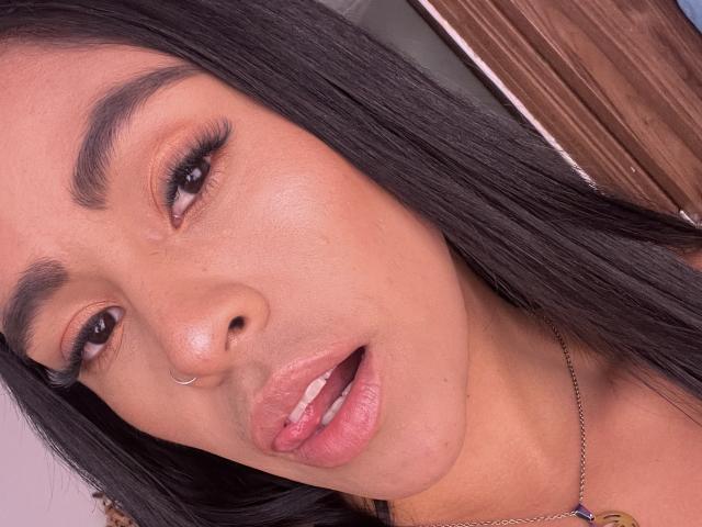 StellaBeckker has tan skin, black straight hair, brown eyes, and a silver nose ring. Her glossy pink lips are slightly open, showing her white teeth.