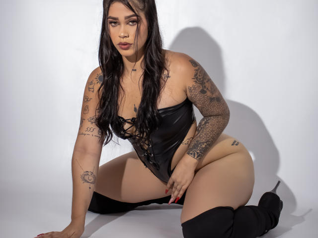 AgathaVelez69 with tattoos, black leather corset, and thigh-high boots, kneeling, showing her ass and pussy, long black hair, serious expression, in a studio with white background.