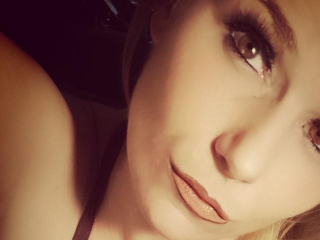 El rostro de DevilsAngel en un selfie, mostrando sus ojos marrones, labios carnosos con un labial rosado y cabello rubio ligeramente ondulado. Su ojo derecho tiene una pequeña cicatriz visible..