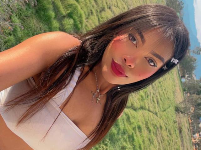 LexieBloom with long brown hair, white crop top, red lipstick, and a cross necklace, showing her tits in a sunny, grassy landscape. She has a cute hair clip.