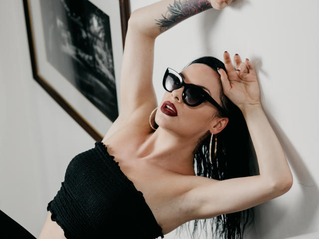 MistressLilyMaria with black tube top, large black sunglasses, red lipstick, and hoop earrings, arms raised, showing armpit tattoo, and long black hair, looking sexy and confident.