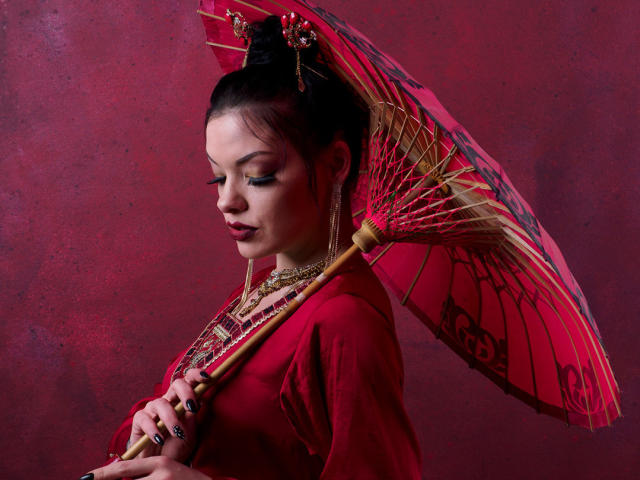 MistressLilyMaria with dark hair in an updo, wearing red clothes, holding a red and gold umbrella, wearing gold jewelry, looking down with closed eyes, red background.