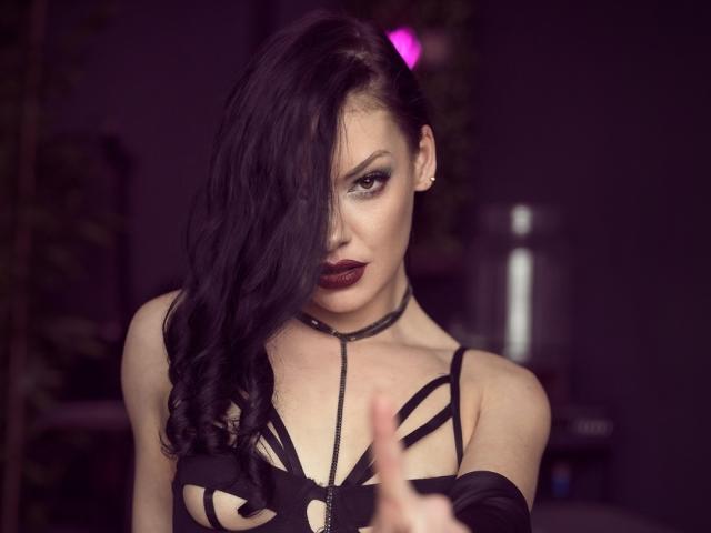 MistressLilyMaria with long black hair, dark makeup, and deep red lipstick, wearing a black strappy top, pointing forward, showing off her tits with a black choker.