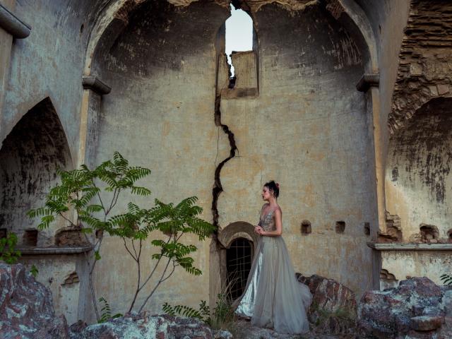 MistressLilyMaria stands in a crumbling, stone archway with a large crack in the wall. She's wearing a flowing, beige dress, and has a tattoo on her right arm.