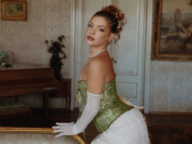 MistressLilyMaria with green lace corset, white gloves, and pearl necklace, showing off her tits and ass, leaning on a vintage couch, in a classical, elegant room.
