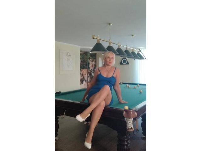 AliceHottie69 mit blonden Haaren, in einem blauen Kleid, mit weißen Schuhen, sitzt auf einem Billardtisch, die Beine gekreuzt, und zeigt ihr Gesäß, in einem Raum mit hängenden Lichtern und Billardkugeln..