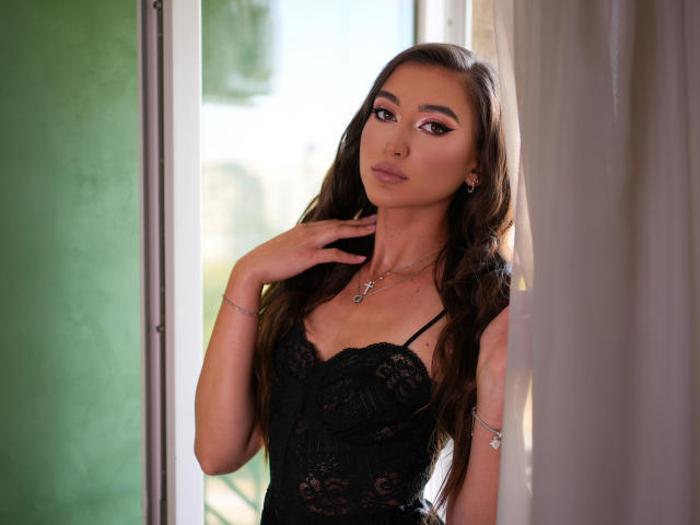 BarbaraLustt with long brown hair, wearing a black lace lingerie top, looking at the camera with her hand near her neck, has a sexy, attractive face, and a small cross necklace.