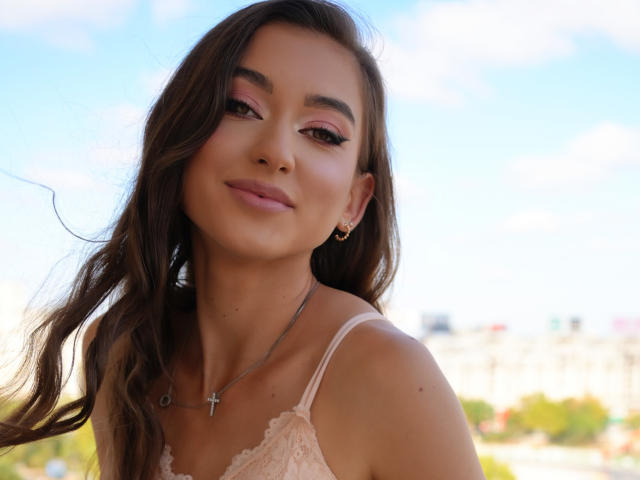BarbaraLustt with long brown hair, pink eyeshadow, subtle makeup, and a cross necklace, smiling softly at the camera, wearing a pink lace top, outside on a sunny day.