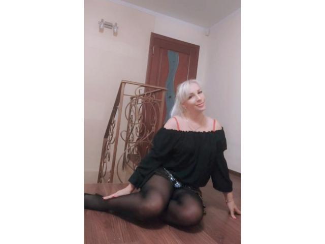 AliceHottie69, com cabelo loiro platinado, sentada no chão de madeira, vestindo uma blusa preta com um ombro descoberto, shorts pretos e meias pretas, sorrindo para a câmera, com uma porta de madeira ao fundo..