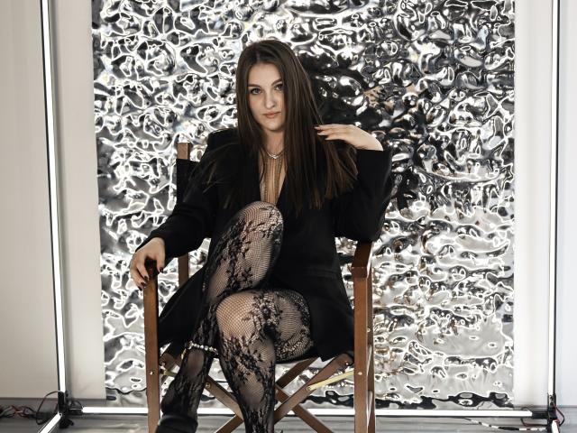 EmmyCharm sits in a wooden director's chair, arms resting on the armrests. She has long brown hair, a serious expression, and wears a black coat over a deep V-neck, revealing a hint of cleavage. Her lace-patterned fishnet stockings are black with floral designs. The background is a textured, metallic, abstract pattern. She is centered, legs crossed, with one hand touching her hair. The image has a professional, studio-lit feel.