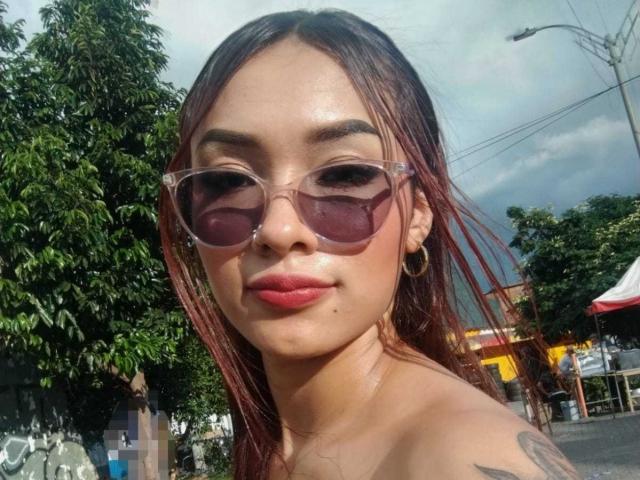 SashaHans, con i capelli tinti di rosso, indossa occhiali da sole trasparenti, rossetto rosa, orecchini a cerchio e un tatuaggio sulla spalla, e guarda la fotocamera con un leggero sorriso..