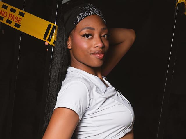 ElisabettaMonroy with dark skin, long black hair, and a headband, wearing a white shirt, her arm raised, showing her tits, smiling with a confident look.