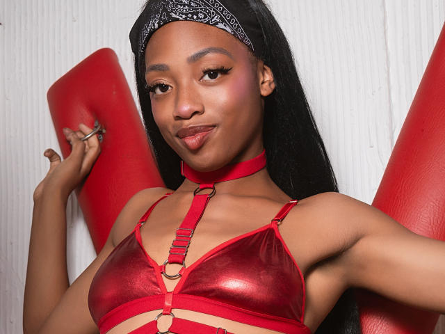 ElisabettaMonroy with dark skin, long black hair, wearing a red shiny bra with straps, black bandana, and red choker, smiling with raised arms, holding red blocks.
