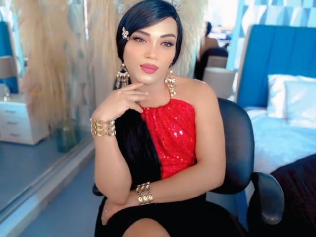 CharlotteSweetX with black hair, red and black sequined top, gold jewelry, and a sexy, confident look, sitting in a chair, her hand on her chin, in a bedroom.