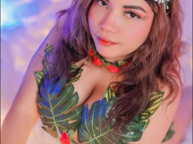 CutieCurvyForeign with green leaf bikini top, red berries, and wavy brown hair, showing off her big boobs and pink lip ring, looking up with a cute smile.