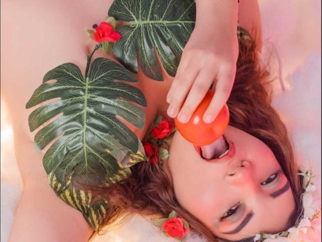 CutieCurvyForeign avec un godemichet rouge-orange dans la bouche, allongée avec de grandes feuilles vertes et des fleurs rouges. Elle a les cheveux bruns bouclés, une peau claire et un piercing au nez..