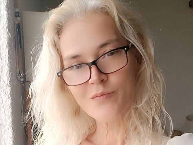 JuliaBlondAngel with blonde, wavy hair, wearing black glasses, has a slight smile, fair skin, and a cute, relaxed face, standing near a window with soft light.