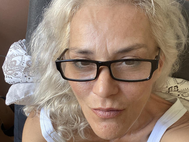 JuliaBlondAngel with pale skin, curly white hair, black rectangular glasses, and a serious expression, wearing a white tank top, showing her slightly wrinkled face and pouty lips.