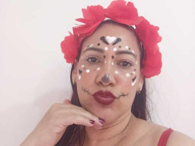 RubieBlaze with painted face, red flower headband, dark lipstick, white and black face paint, heart on forehead, puffy red lips, puffy nose, hand on chin, red nail polish.