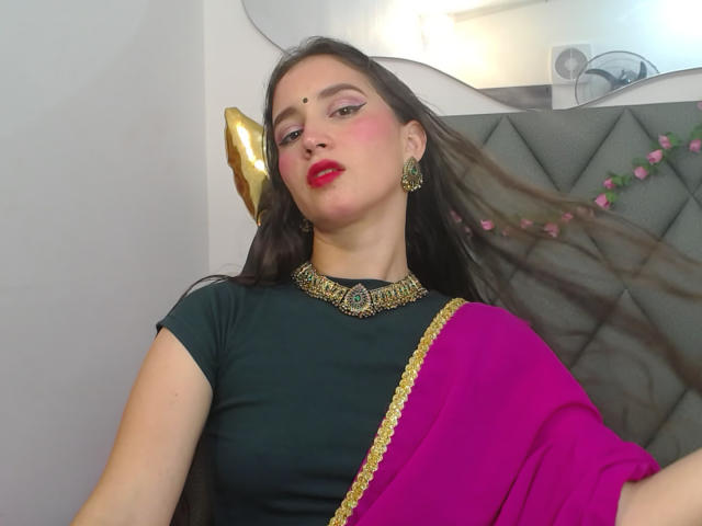 JalimaMona with long brown hair, red lipstick, green top, pink sari, gold jewelry, and a nose ring, sitting, looking at camera, with a confident, slightly smug expression.