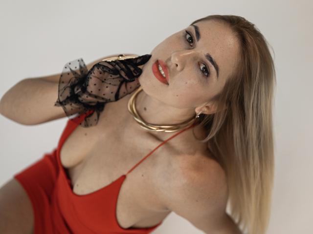 ArishkaLulu tem cabelo loiro, um vestido vermelho que revela seus seios com um decote baixo, está usando uma luva preta com bolinhas, um colar de ouro e batom vermelho, olhando para cima com um sorriso sedutor..