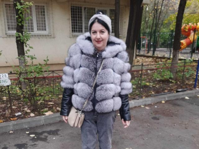 LolaGreate wearing a puffy gray fur vest over a black leather jacket, gray pants, and a gray beanie, smiling while holding a beige purse, standing on a sidewalk.