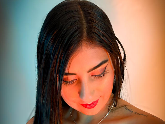 PaullieEvans has black hair, red lipstick, and a tattoo on her shoulder. She's looking down with her eyes closed, wearing a silver necklace, and has thick eyebrows.