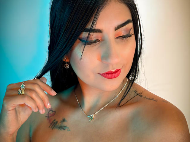 PaullieEvans with black hair, red lipstick, and tattoos, wearing a necklace and ring, holding her hair, showing her tits, and a smile, against a blue and white background.