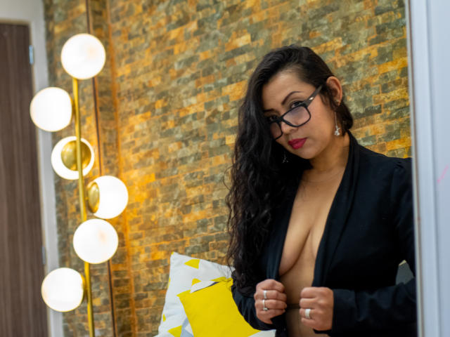 ThaliaSaenz stands in a modern room with a textured stone wall. She has long, wavy black hair and wears glasses with a confident expression. Her black blazer is open, revealing large, natural breasts. She holds a yellow file and a white, geometrically-patterned sheet. A tall, modern floor lamp with spherical white bulbs is to her left. Her red lipstick contrasts with her dark hair and blazer. She has a ring on her right hand. The image is sharp and well-lit.