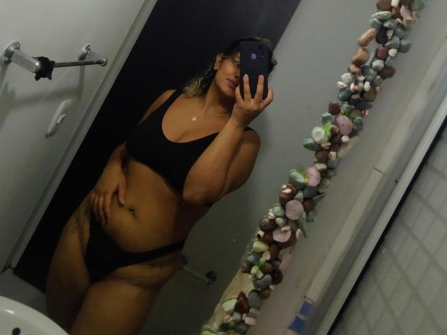 MariGonz with tan skin, black bra, and black panties, holding phone in mirror selfie, showing tits and ass, standing in bathroom with stone-decorated mirror.
