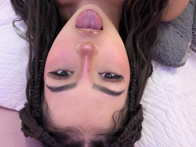 RheaVolk's face upside down, tongue out, with pink lips, brown eyes, and dark braided hair, lying on a white quilted surface, looking playful. Her nose is slightly upturned.