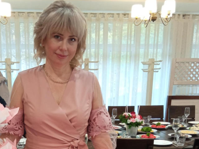 LaurenWishy, an older woman with short, wavy blonde hair, wears a pink, satin, V-neck dress with lace sleeves. She has a small smile and is seated at a dining table. The table has clear glasses, plates, and a small bouquet of pink flowers. She wears a gold necklace. The background features white curtains and a chandelier. The image is well-lit with natural light. Her skin is fair, and she has blue eyes.