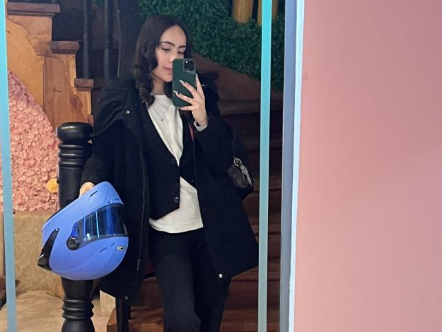 AitanaJohns mit welligem, braunem Haar, einem schwarzen Mantel, einem weißen Hemd und einer schwarzen Hose, die einen blauen Helm hält, macht ein Selfie mit einem Spiegel, ein grünes Telefon in der Hand..
